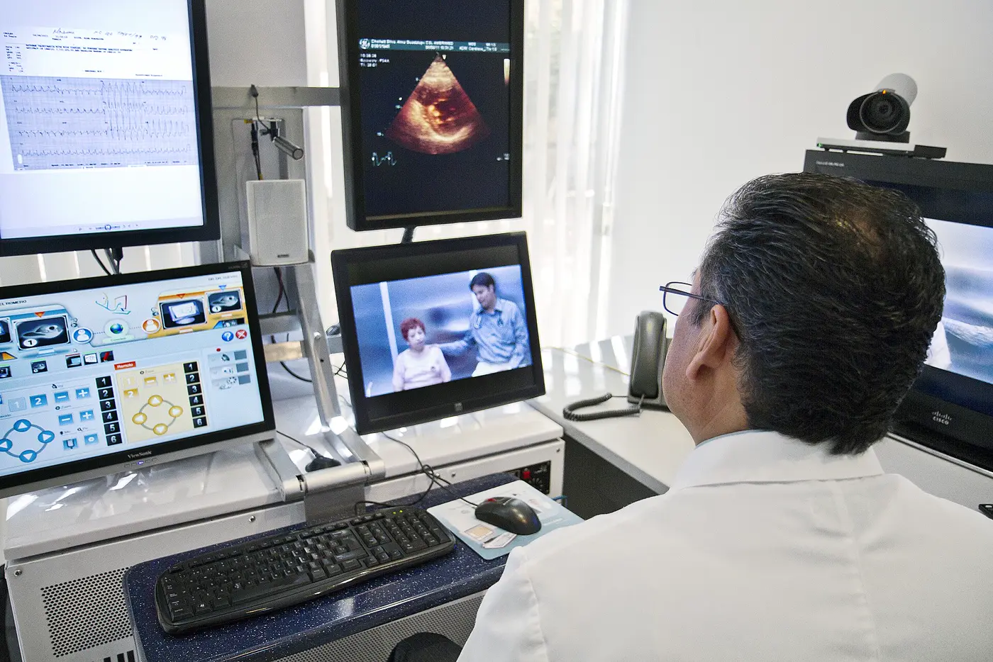 A doctor at a workstation on a live video consultation with a patient, reports and scans on the screens beside him